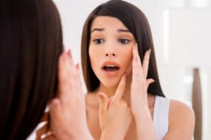 Frustrated young woman examining her face while looking at the mirror